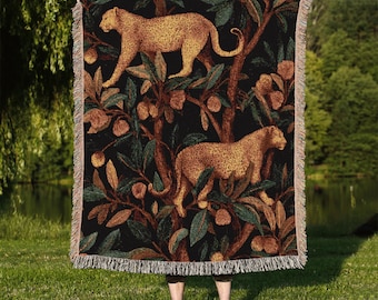 May include: A black woven throw blanket featuring a repeating pattern of two leopards amidst a stylized tree with green leaves and brown fruit. The leopards are tan with dark spots. The blanket has a fringed edge.