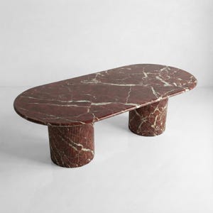 Pode incluir: Uma mesa de centro ovalada em mármore vermelho com veios brancos. A mesa tem duas bases cilíndricas com ranhuras verticais. A mesa tem um design moderno e está sobre um fundo branco.