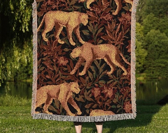 May include: A woven throw blanket featuring a repeating pattern of golden leopards amidst a backdrop of dark brown and green foliage. The blanket has a fringed edge and is held up in front of a person.