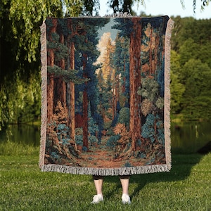 May include: A woven tapestry blanket depicting a forest scene with tall trees in shades of brown, green, and blue. The blanket has a fringed edge. The blanket is held up against a backdrop of green grass and a lake.