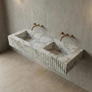 Peut inclure: Un évier mural moderne en marbre avec deux vasques et des robinets dorés. L'évier présente un motif veiné blanc et gris et un extérieur rainuré verticalement. Les robinets sont fixés au mur gris au-dessus de l'évier.