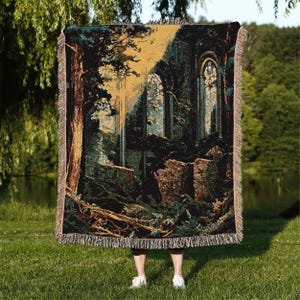 May include: Woven throw blanket featuring a detailed illustration of a forest scene with a stone structure, trees, and sunlight. The blanket has a fringed edge and is primarily in shades of green, brown, and gold. The design evokes a sense of mystery and nature.