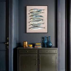 May include: A framed art print featuring a vertical arrangement of seven silver fish with dark eyes and blue-green highlights. The artwork is displayed on a light beige background, set against a dark blue wall. A dark green cabinet with decor is below.