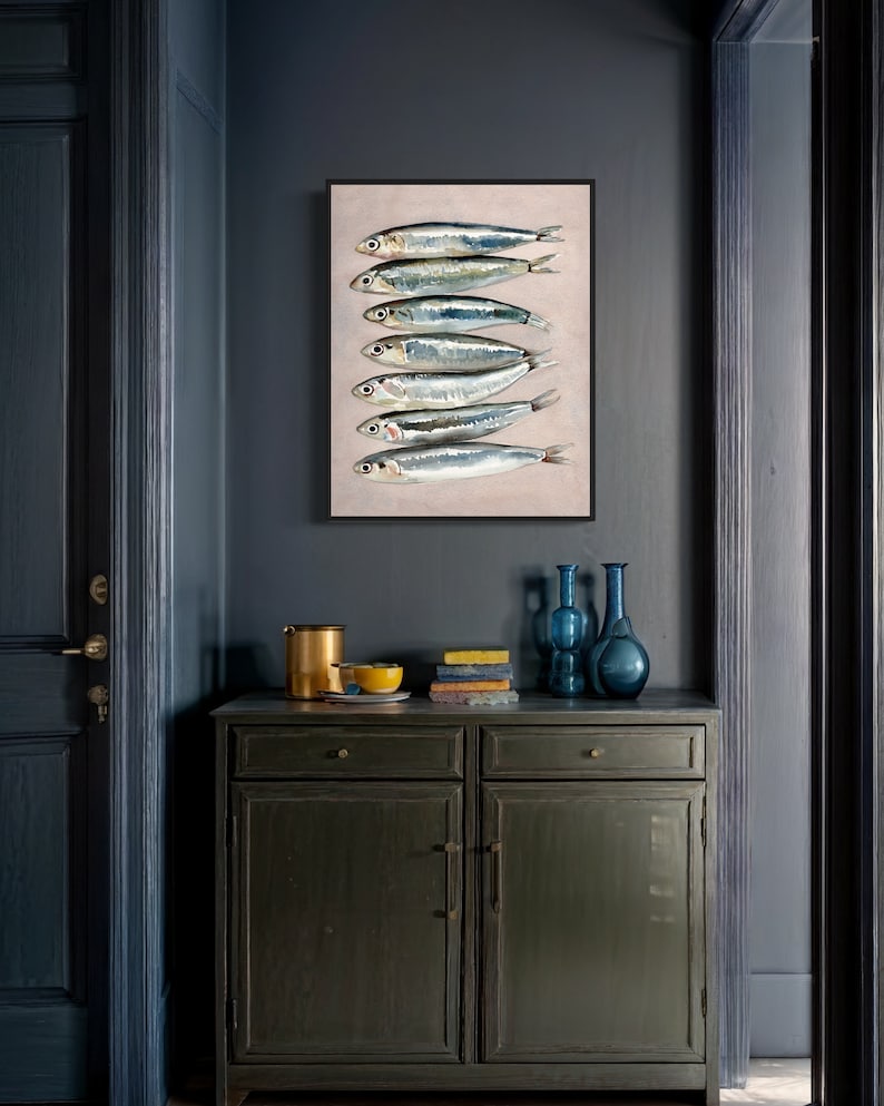 May include: A framed art print featuring a vertical arrangement of seven silver fish with dark eyes and blue-green highlights. The artwork is displayed on a light beige background, set against a dark blue wall. A dark green cabinet with decor is below.