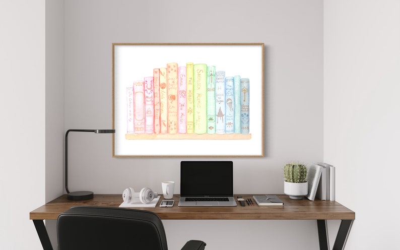 May include: Watercolour print of a collection of colourful books on a shelf. The books are in shades of pink, yellow, green, and blue. The artwork is framed in a light brown frame and hangs on a grey wall above a wooden desk.