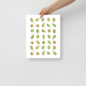 May include: A white print featuring a watercolor illustration of green olives with red centers and small green leaves. The olives are scattered across the print. The print is held by a person's hand.