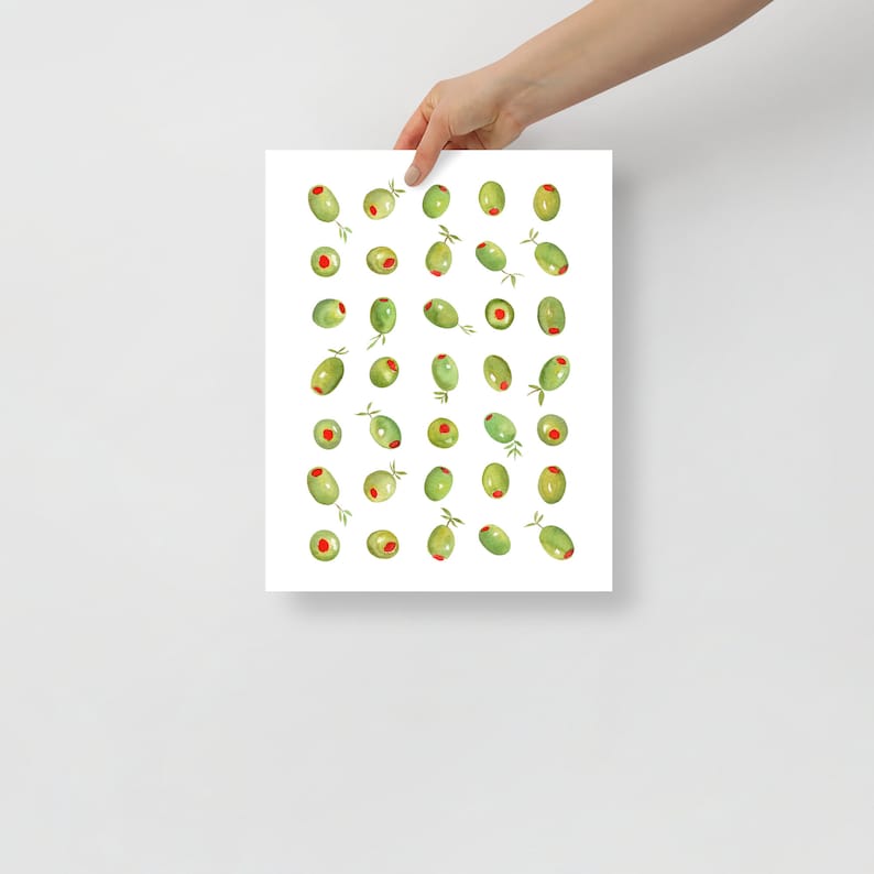 May include: A white print featuring a watercolor illustration of green olives with red centers and small green leaves. The olives are scattered across the print. The print is held by a person's hand.