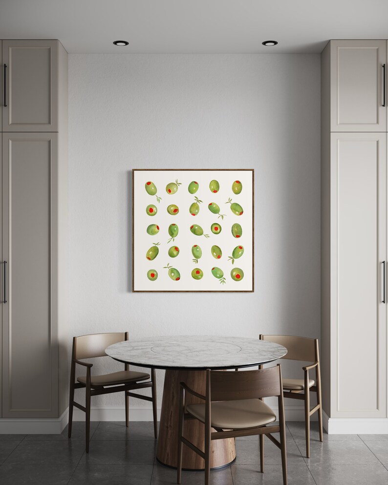 May include: A framed print featuring a watercolor illustration of green olives with red pimentos. The artwork is displayed on a white wall above a round marble-topped table with wooden chairs. The room has neutral-toned cabinets.