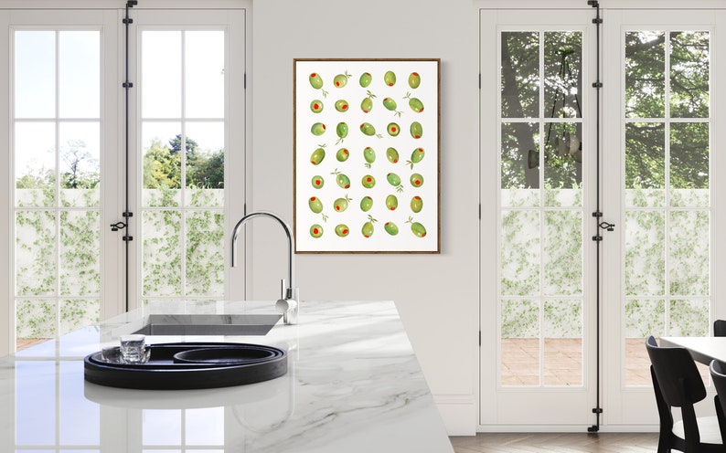 May include: A framed print featuring a pattern of green olives with red centers and small green leaves. The artwork is displayed in a bright kitchen setting with white walls, a marble countertop, and large windows.