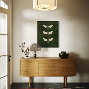 May include: A rectangular wall art print featuring three detailed illustrations of bees arranged vertically against a dark forest green background, enclosed in a thin black frame, displayed above a light-toned wooden sideboard cabinet.