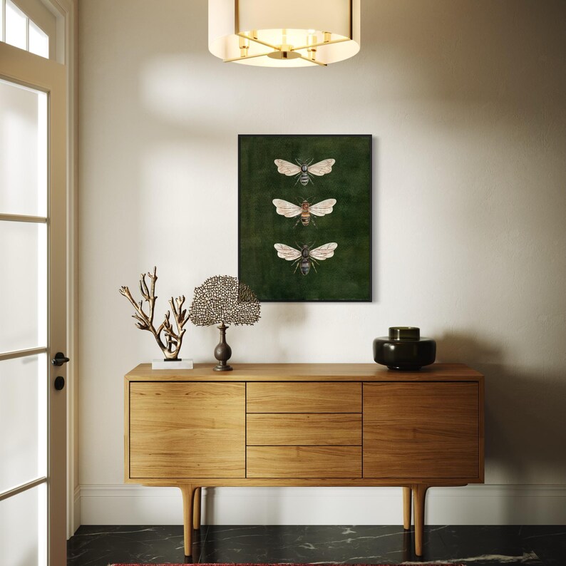 May include: A rectangular wall art print featuring three detailed illustrations of bees arranged vertically against a dark forest green background, enclosed in a thin black frame, displayed above a light-toned wooden sideboard cabinet.