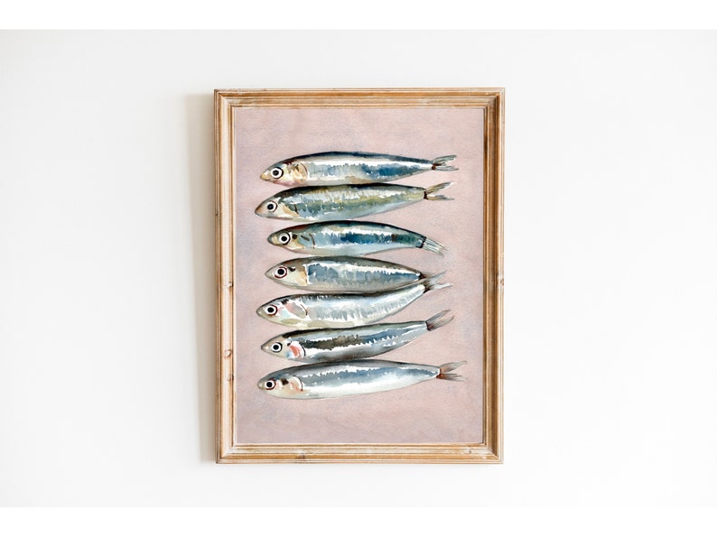 May include: Watercolor print of seven sardines arranged vertically on a light pink background. The fish are silver with dark eyes and fins, and the artwork is framed in a light brown wooden frame. A decorative piece for a kitchen or dining area.