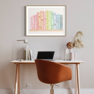 May include: A framed watercolour print featuring a collection of colourful books. The books are arranged on a wooden shelf, with titles visible. The artwork is displayed above a desk with a laptop, chair, and decorative items.