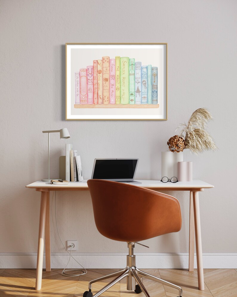 May include: A framed watercolour print featuring a collection of colourful books. The books are arranged on a wooden shelf, with titles visible. The artwork is displayed above a desk with a laptop, chair, and decorative items.