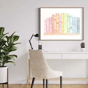 May include: Watercolour print of a collection of colourful books on a shelf, framed in a light brown wooden frame. The books are in shades of pink, yellow, green, and blue. The artwork is displayed on a white desk with a black desk lamp and a potted plant.