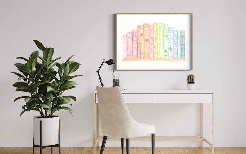 May include: Watercolour print of a collection of colourful books on a shelf, framed in a light brown wooden frame. The books are in shades of pink, yellow, green, and blue. The artwork is displayed on a white desk with a black desk lamp and a potted plant.