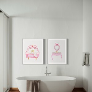 May include: Two framed watercolor illustrations of a pink bathtub and a vanity set with a mirror, both with pink bows. The bathtub has gold accents and a white towel. The vanity set has a pink countertop and gold legs. The illustrations are hanging on a white wall in a bathroom with a white bathtub.