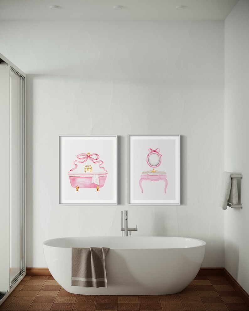 May include: Two framed watercolor illustrations of a pink bathtub and a vanity set with a mirror, both with pink bows. The bathtub has gold accents and a white towel. The vanity set has a pink countertop and gold legs. The illustrations are hanging on a white wall in a bathroom with a white bathtub.