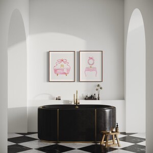 May include: A black freestanding bathtub with gold trim sits on a black and white checkered floor. Two framed watercolor prints of a pink bathtub and a vanity with a mirror hang on the wall above the bathtub.