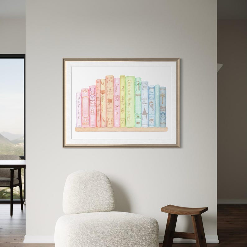 May include: A framed watercolour print featuring a collection of colourful books on a wooden shelf. The books are in shades of pink, orange, yellow, green, and blue. The print is displayed on a light grey wall, with a white chair and wooden stool in the foreground.