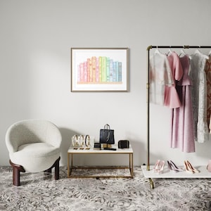 May include: A framed watercolour print depicting a row of colourful books. The books are in shades of pink, yellow, green, and blue. The print is displayed on a white wall above a small table with accessories and a chair.
