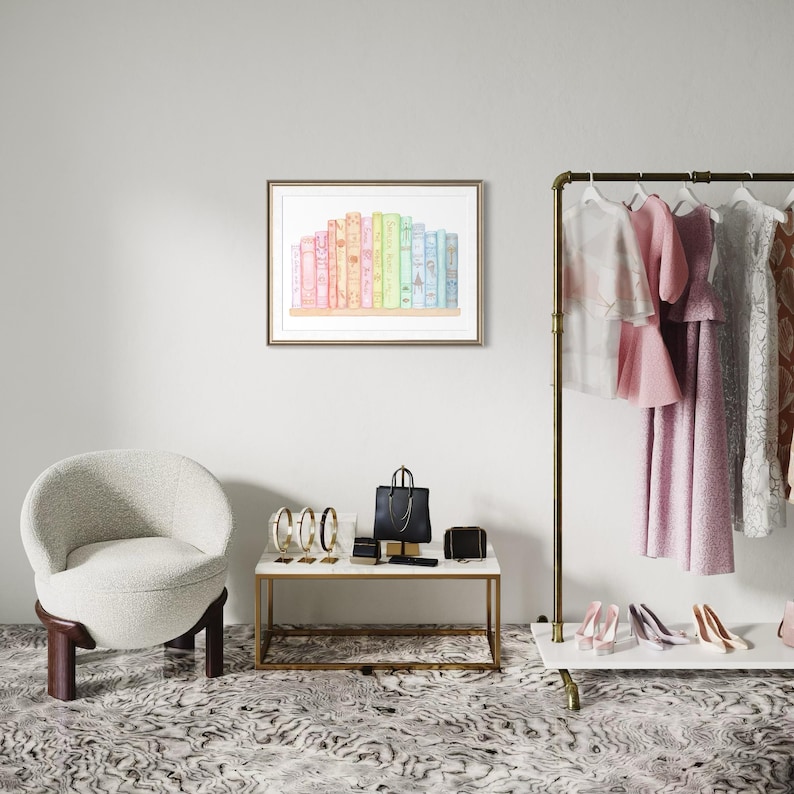 May include: A framed watercolour print depicting a row of colourful books. The books are in shades of pink, yellow, green, and blue. The print is displayed on a white wall above a small table with accessories and a chair.