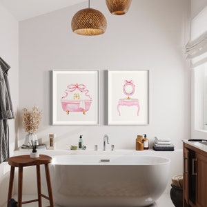 May include: Two framed watercolor illustrations of a pink bathtub with a bow and a pink vanity with a mirror and a bow. The illustrations are hanging on a white wall in a bathroom with a white bathtub, a wooden stool, and a brown cabinet.