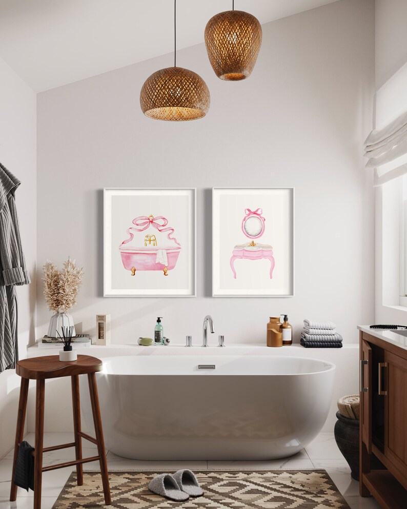 May include: Two framed watercolor illustrations of a pink bathtub with a bow and a pink vanity with a mirror and a bow. The illustrations are hanging on a white wall in a bathroom with a white bathtub, a wooden stool, and a brown cabinet.