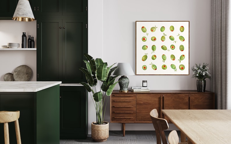 May include: A framed watercolor print featuring green olives with red centers and small sprigs. The artwork hangs above a wooden sideboard in a dining room setting. A large potted plant and a table are also visible.