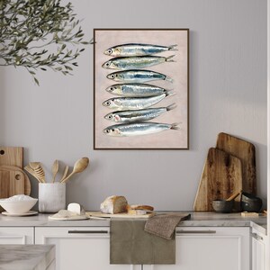 May include: A framed art print featuring a vertical arrangement of seven sardines. The fish are silver with dark stripes, set against a light pink background. The artwork is displayed in a kitchen setting with wooden cutting boards and utensils.