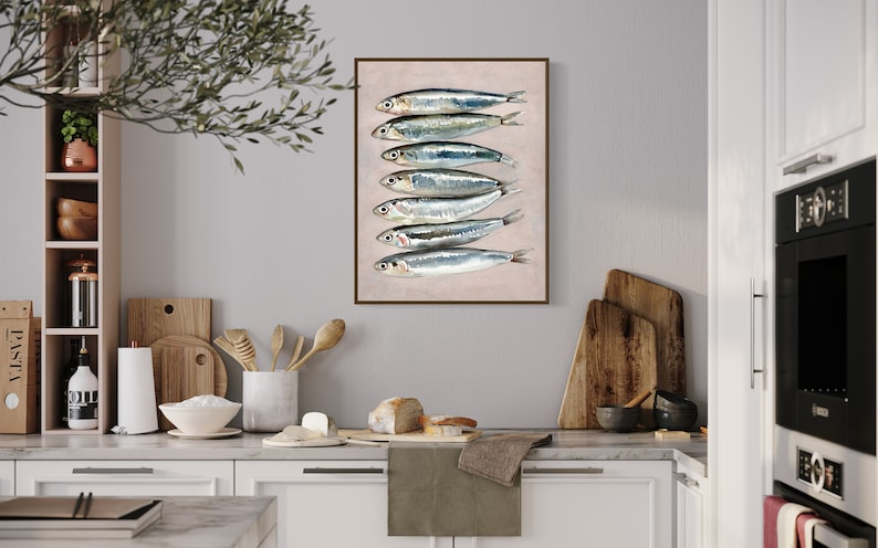 May include: A framed art print featuring a vertical arrangement of seven sardines. The fish are silver with dark stripes, set against a light pink background. The artwork is displayed in a kitchen setting with wooden cutting boards and utensils.