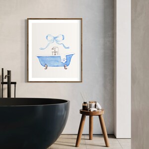 Peut inclure: Une peinture &agrave; l'aquarelle d'une baignoire bleue avec une serviette blanche et un n&oelig;ud bleu sur le dessus. La baignoire a quatre pieds et un robinet. La peinture est encadr&eacute;e dans un cadre brun.