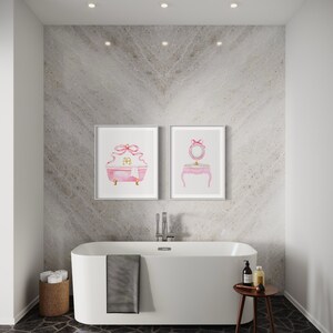 May include: A white bathtub with a gray towel hanging over the side, a wooden stool with a glass jar and bottles on top, and two framed watercolor prints of pink vanity sets with gold accents on a gray wall.