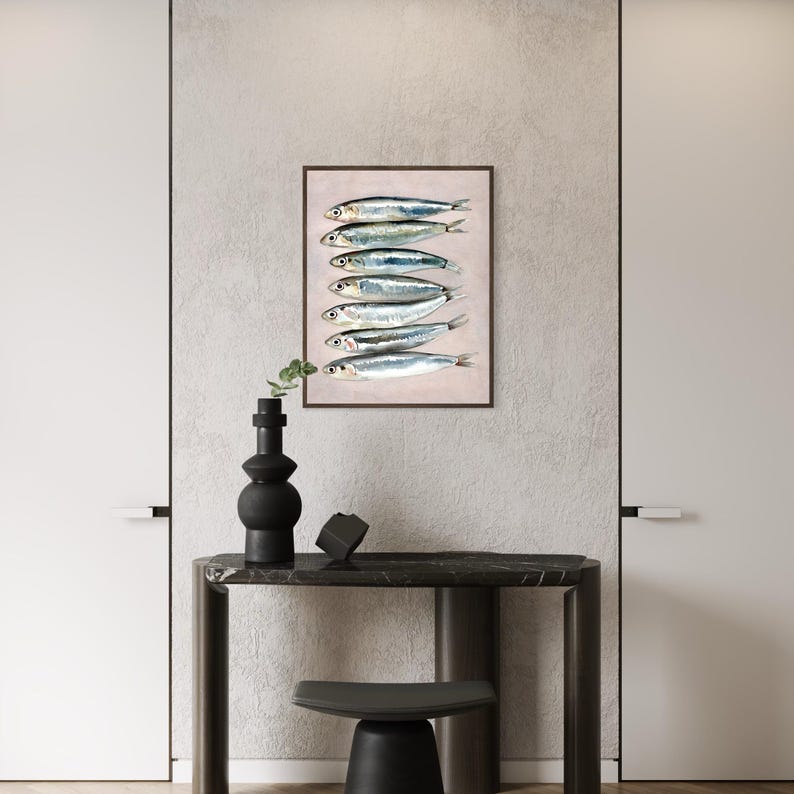 May include: A framed art print featuring a watercolor illustration of seven sardines arranged vertically. The fish are silver with dark stripes and black eyes, set against a pale pink background. The frame is dark brown. The artwork is displayed on a wall.