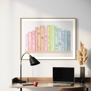May include: A framed watercolour print featuring a collection of books with titles like "Sherlock Holmes" and "The Hobbit." The artwork, in a gold frame, sits above a desk with a black lamp, laptop, and a red apple.