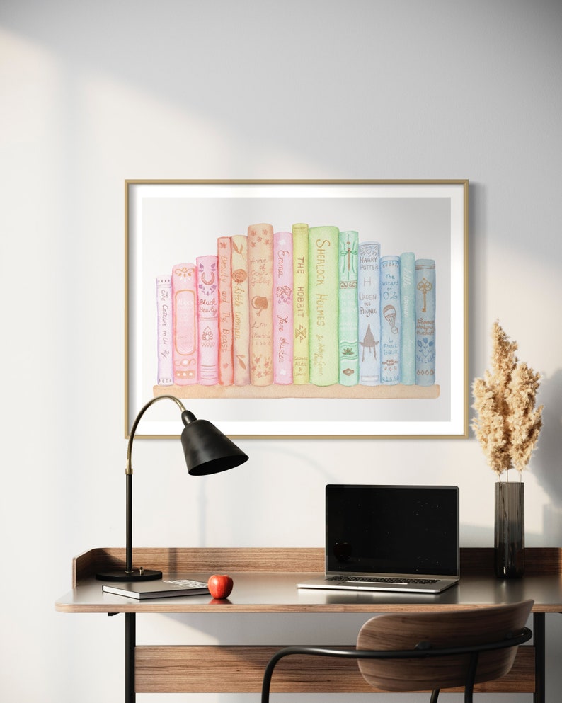 May include: A framed watercolour print featuring a collection of books with titles like "Sherlock Holmes" and "The Hobbit." The artwork, in a gold frame, sits above a desk with a black lamp, laptop, and a red apple.