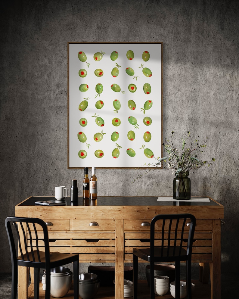 May include: A framed print featuring a watercolor illustration of green olives with red centers and small green leaves. The artwork hangs above a wooden cabinet with black chairs, bottles, and a vase.