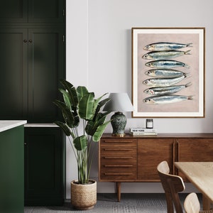 May include: A framed art print featuring a stack of sardines. The fish are silver with blue and black markings, set against a light pink background. The artwork is displayed in a room with dark green cabinets, a wooden sideboard, and a large potted plant.