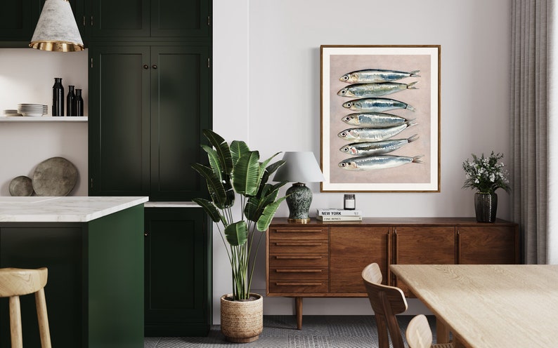 May include: A framed art print featuring a stack of sardines. The fish are silver with blue and black markings, set against a light pink background. The artwork is displayed in a room with dark green cabinets, a wooden sideboard, and a large potted plant.