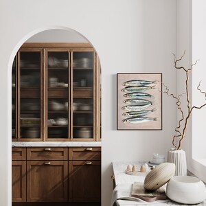 May include: A framed print of sardines hangs in a kitchen setting. The artwork features seven fish with silver and blue scales on a beige background. The kitchen has wooden cabinets, a marble countertop, and a window with a view of greenery.