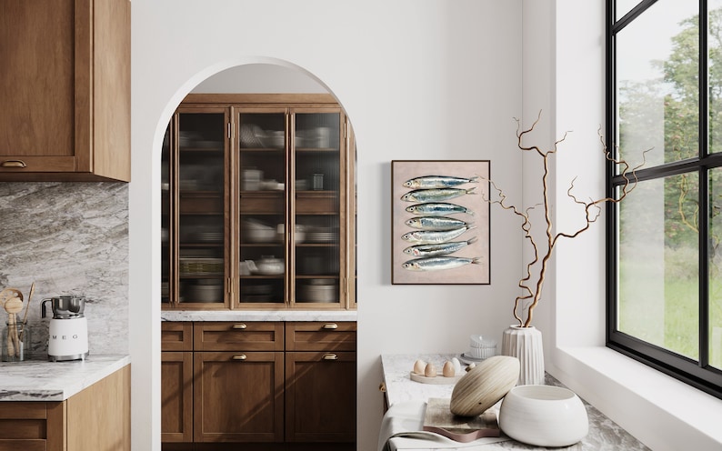May include: A framed print of sardines hangs in a kitchen setting. The artwork features seven fish with silver and blue scales on a beige background. The kitchen has wooden cabinets, a marble countertop, and a window with a view of greenery.