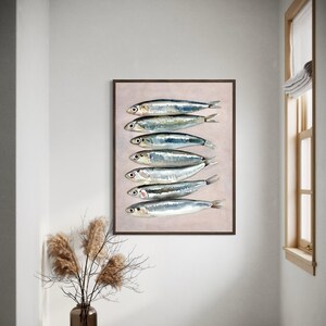 May include: A framed art print featuring a watercolor painting of seven sardines arranged vertically. The fish are silver with dark eyes and fins, set against a pale pink background. The frame is dark brown.