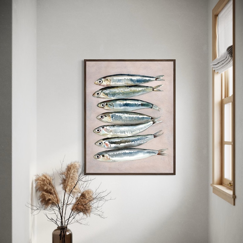May include: A framed art print featuring a watercolor painting of seven sardines arranged vertically. The fish are silver with dark eyes and fins, set against a pale pink background. The frame is dark brown.