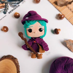 May include: A handmade crochet doll of a witch with teal hair, a purple hat and cloak, and brown boots. The doll is holding a brown staff and has a small brown bag. The doll is surrounded by small wooden mushrooms and a ball of purple yarn.