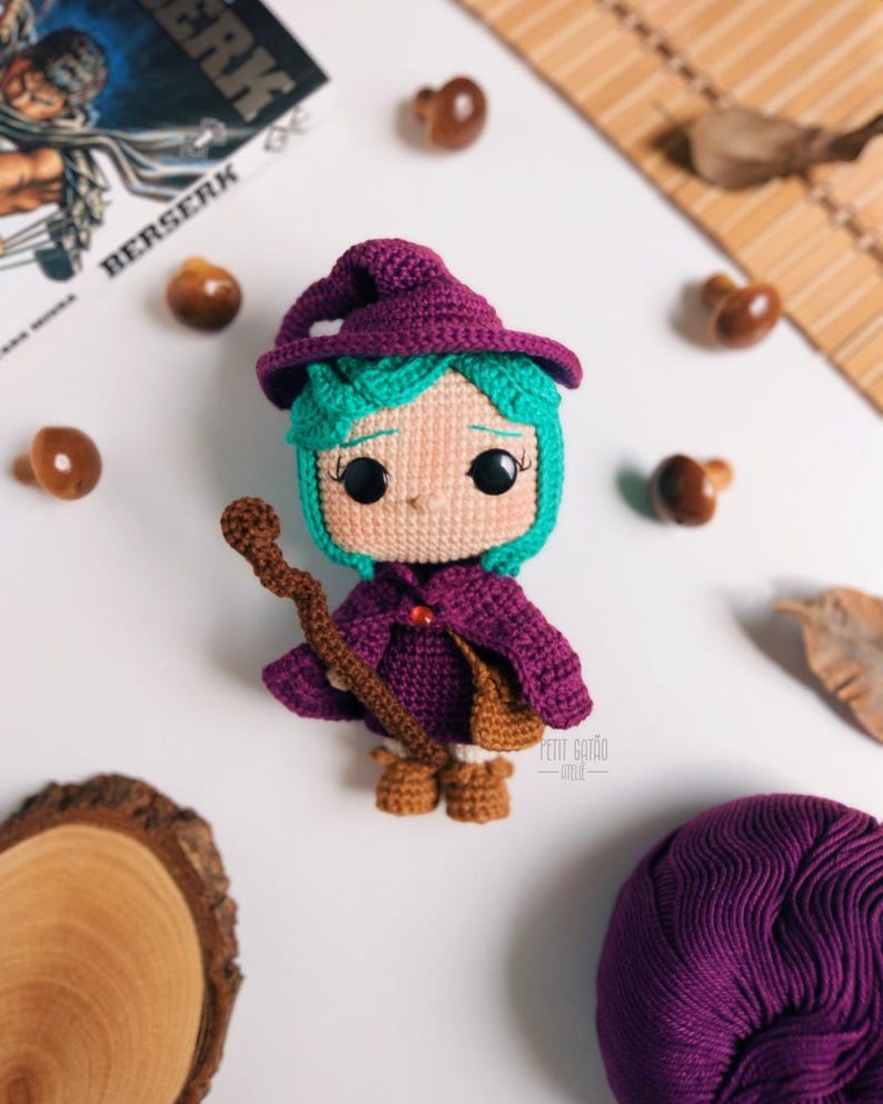 May include: A handmade crochet doll of a witch with teal hair, a purple hat and cloak, and brown boots. The doll is holding a brown staff and has a small brown bag. The doll is surrounded by small wooden mushrooms and a ball of purple yarn.