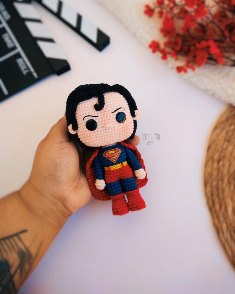Op de afbeelding: Een handgemaakte gehaakte Superman-pop met een rode cape, blauw en rood pak en zwart haar. De pop heeft het Superman-logo op de borst. De pop wordt in een hand gehouden tegen een witte achtergrond.