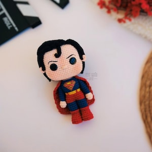 Op de afbeelding: Een handgemaakte gehaakte Superman-pop. De pop heeft zwart haar, blauwe ogen en een rood en blauw kostuum met het Superman-logo. De pop heeft een rode cape en rode laarzen. De pop ligt op een wit oppervlak.