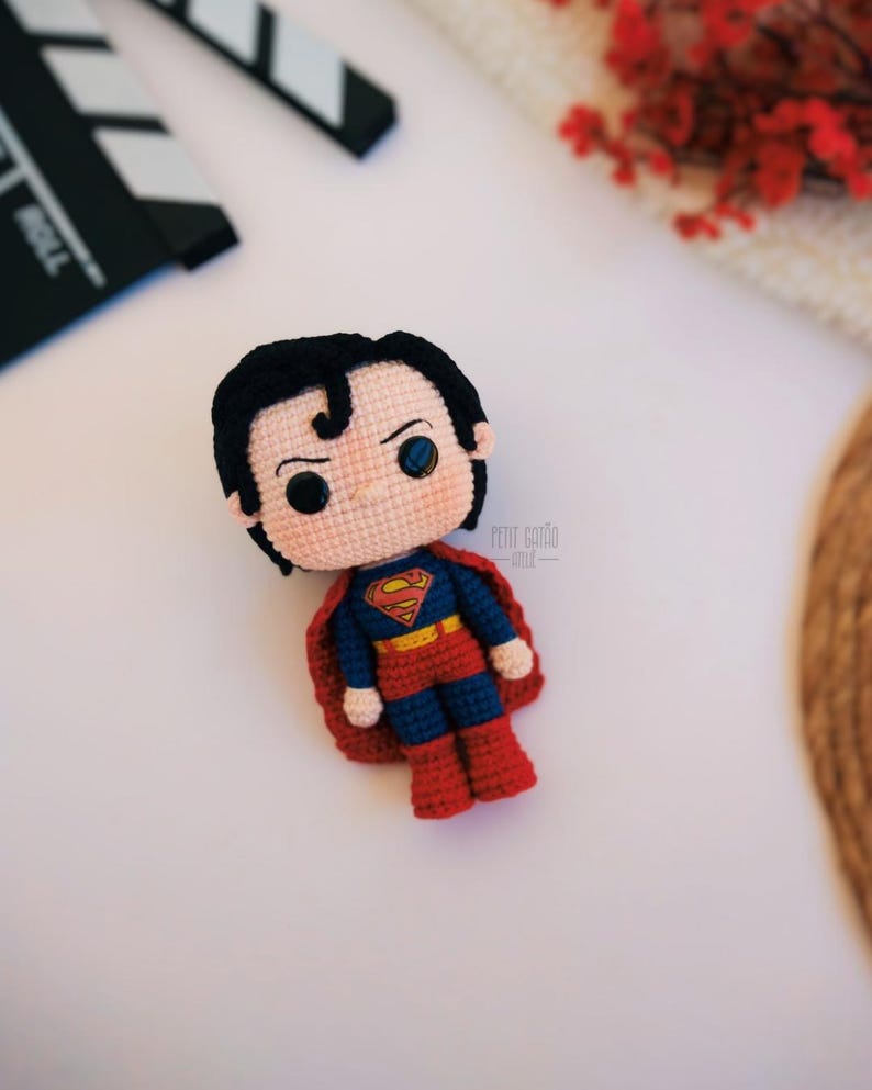 Op de afbeelding: Een handgemaakte gehaakte Superman-pop. De pop heeft zwart haar, blauwe ogen en een rood en blauw kostuum met het Superman-logo. De pop heeft een rode cape en rode laarzen. De pop ligt op een wit oppervlak.