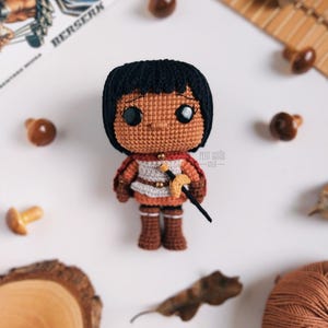 May include: A handmade crochet doll with black hair, brown skin, and a warrior outfit. The doll is holding a small sword and wearing a red cape. The doll is surrounded by decorative mushrooms and a wooden slice.