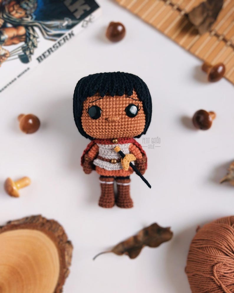 May include: A handmade crochet doll with black hair, brown skin, and a warrior outfit. The doll is holding a small sword and wearing a red cape. The doll is surrounded by decorative mushrooms and a wooden slice.